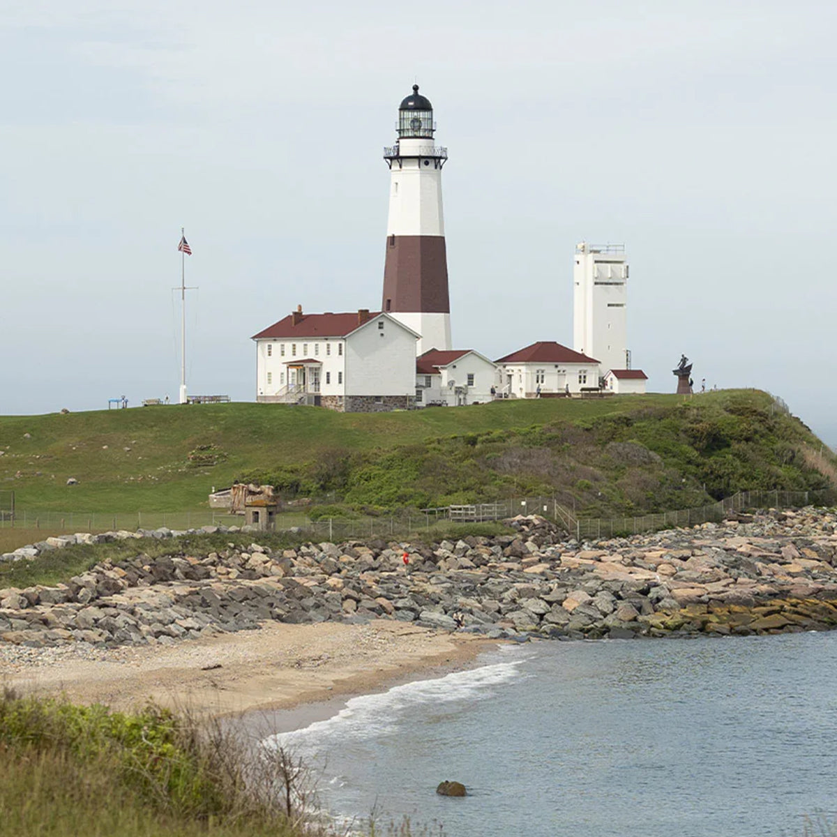 A coastal lighthouse with red-roofed buildings, grassy hill, and rocky shoreline along the ocean.