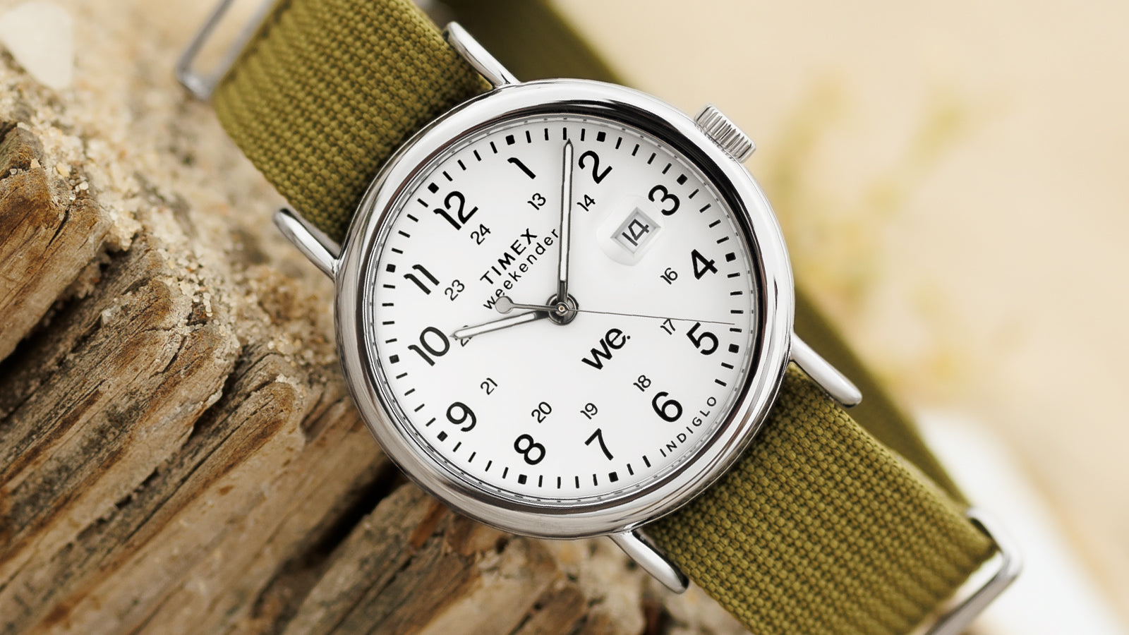 Timex Weekender watch with a green fabric strap resting on driftwood, showing the white dial, black numerals, and date display.