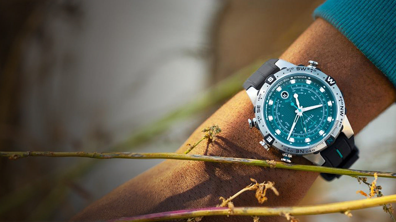 A Timex Expedition Tide-Temp-Compass watch with a teal dial and black silicone strap, worn on the wrist in an outdoor setting. The stainless-steel case features a compass bezel with bold cardinal direction markers.