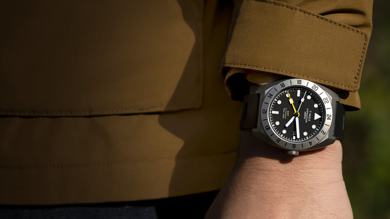 A Timex Expedition North GMT Titanium Automatic watch with a black dial and bold 24-hour bezel, worn on the wrist with a brown outdoor jacket.