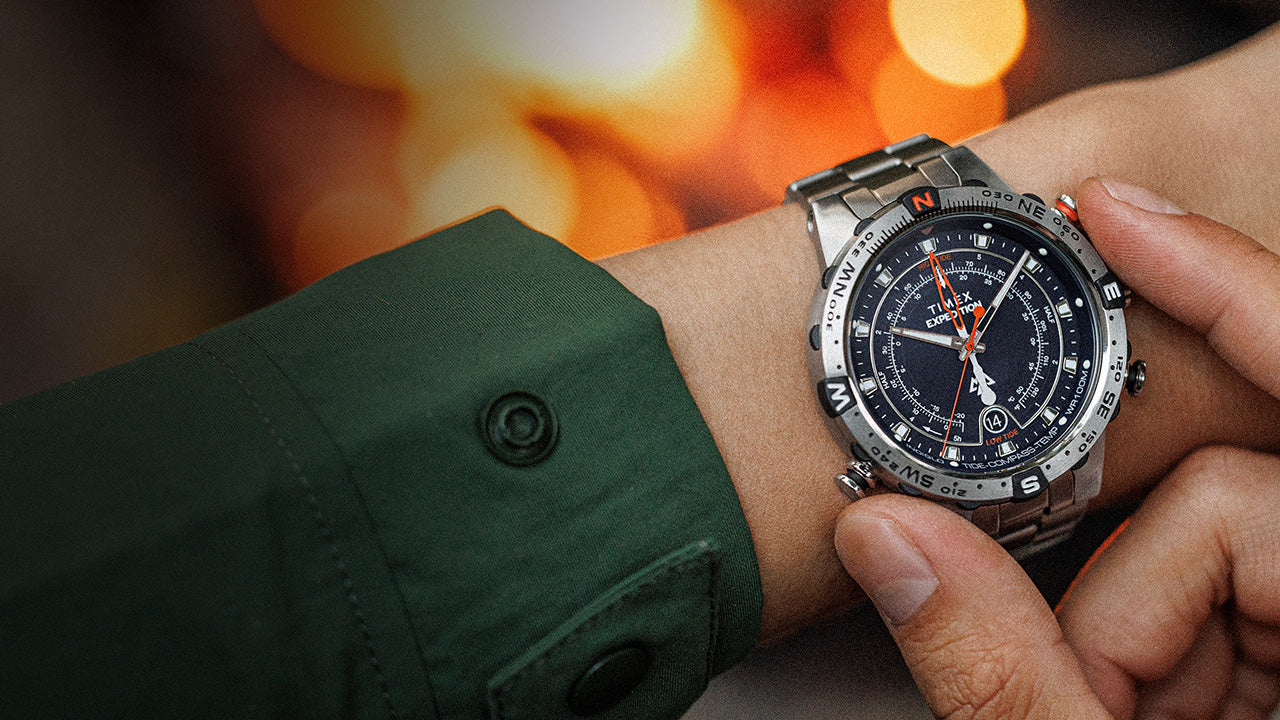 Close-up of a Timex Expedition Tide-Temp-Compass watch on a wrist, with a stainless-steel bracelet and multifunction black dial featuring tide, temperature, and compass indicators. The glowing firelight in the background highlights the rugged, outdoor-ready design.
