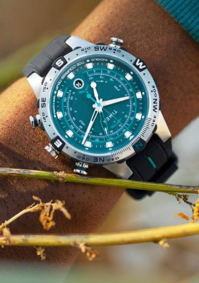 A Timex Expedition Tide-Temp-Compass watch with a teal dial and black silicone strap, worn on the wrist in an outdoor setting. The stainless-steel case features a compass bezel with bold cardinal direction markers.