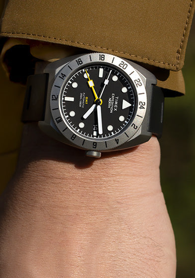 A Timex Expedition North GMT Titanium Automatic watch with a black dial and bold 24-hour bezel, worn on the wrist with a brown outdoor jacket.