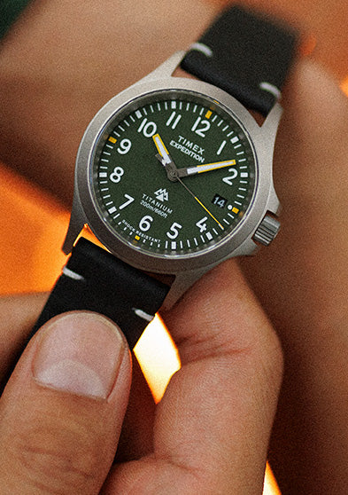 A Timex Expedition Titanium watch with a green dial, luminous white numerals, and black leather strap, worn on the wrist in warm lighting.