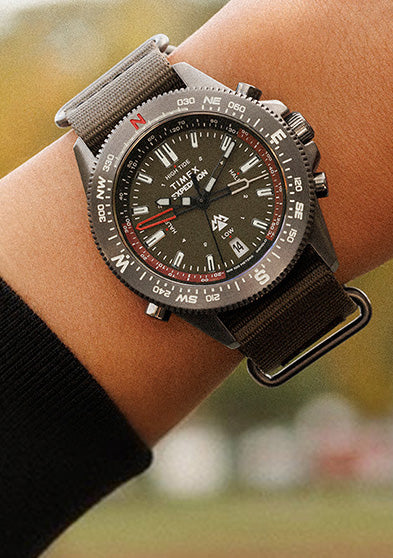 Timex Expedition Tide-Temp-Compass worn on the wrist, featuring a black dial, compass bezel, and green fabric strap, shown against a soft outdoor background.