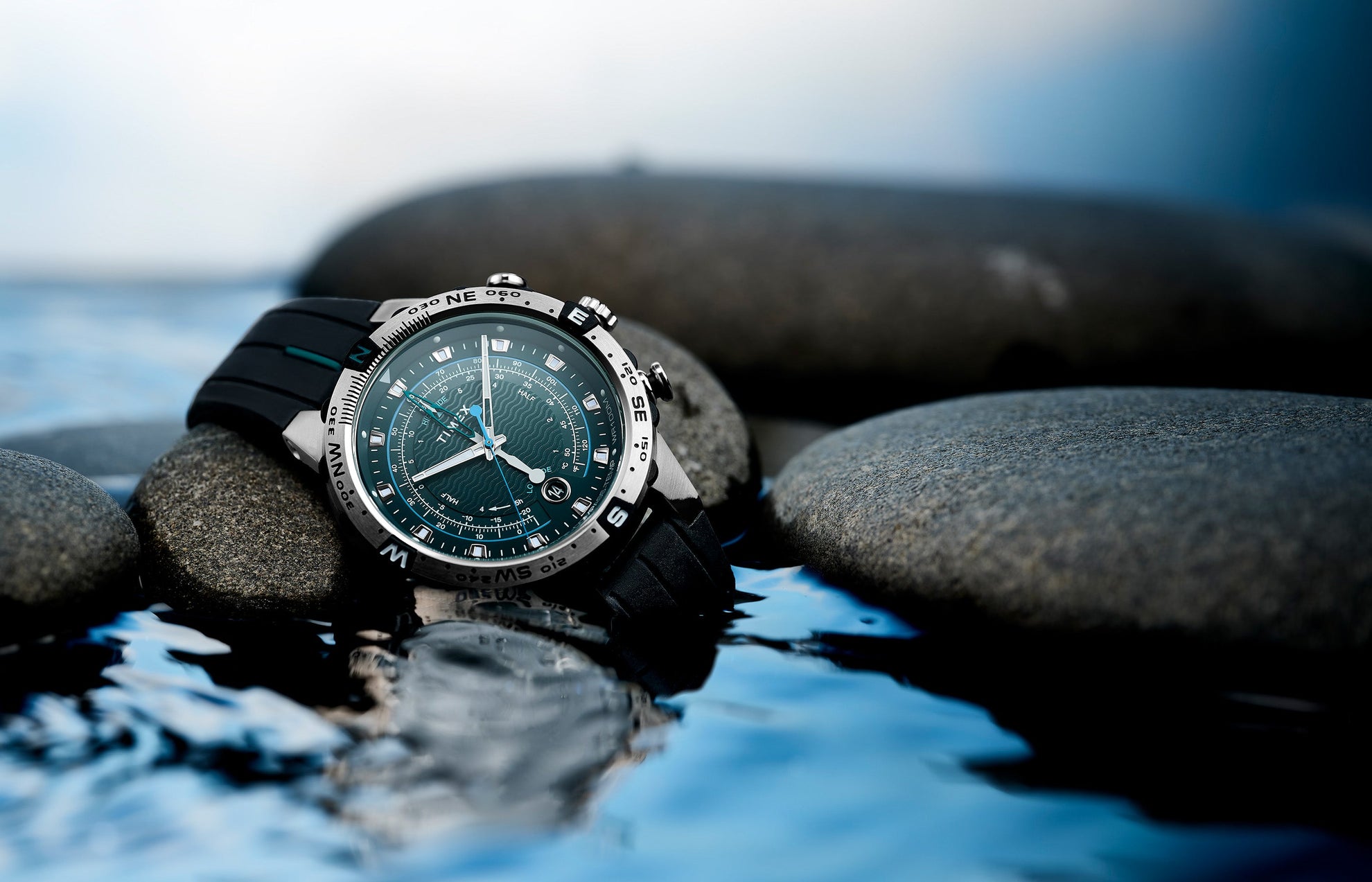 Close-up of the Timex Expedition Tide-Temp-Compass with a teal wave-pattern dial resting on smooth rocks beside water, showcasing its rugged, water-resistant design.