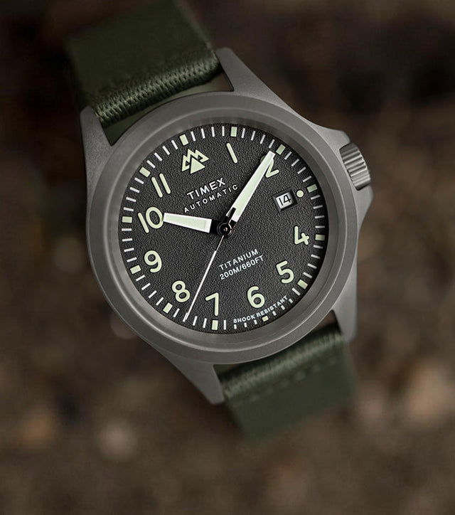 A Timex Expedition North Titanium Automatic watch with a matte titanium case, black textured dial, luminous numerals, and green fabric strap, placed on a rugged surface.