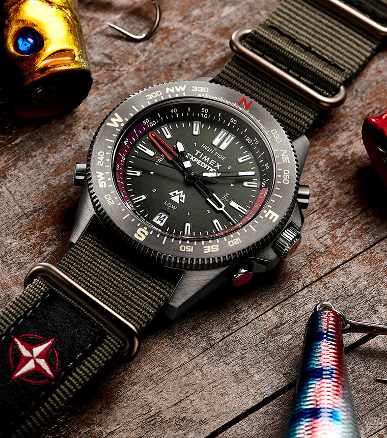 Timex Expedition Tide-Temp-Compass with green fabric strap displayed on a wooden surface among colorful fishing lures, highlighting its rugged outdoor style.