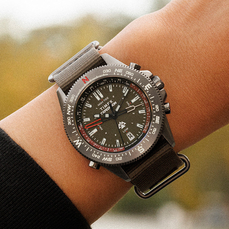 Timex Expedition Tide-Temp-Compass worn on the wrist, featuring a black dial, compass bezel, and green fabric strap, shown against a soft outdoor background.
