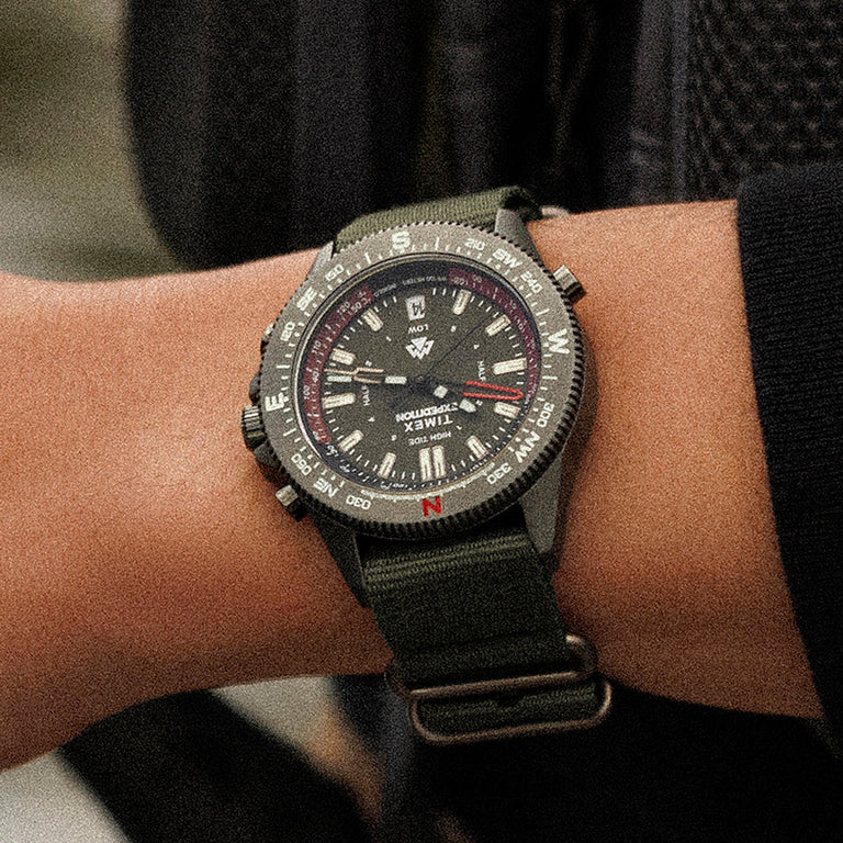 Timex Expedition Tide-Temp-Compass on wrist with green strap and bold black dial, compass bezel with directional markings visible in a darker outdoor setting.