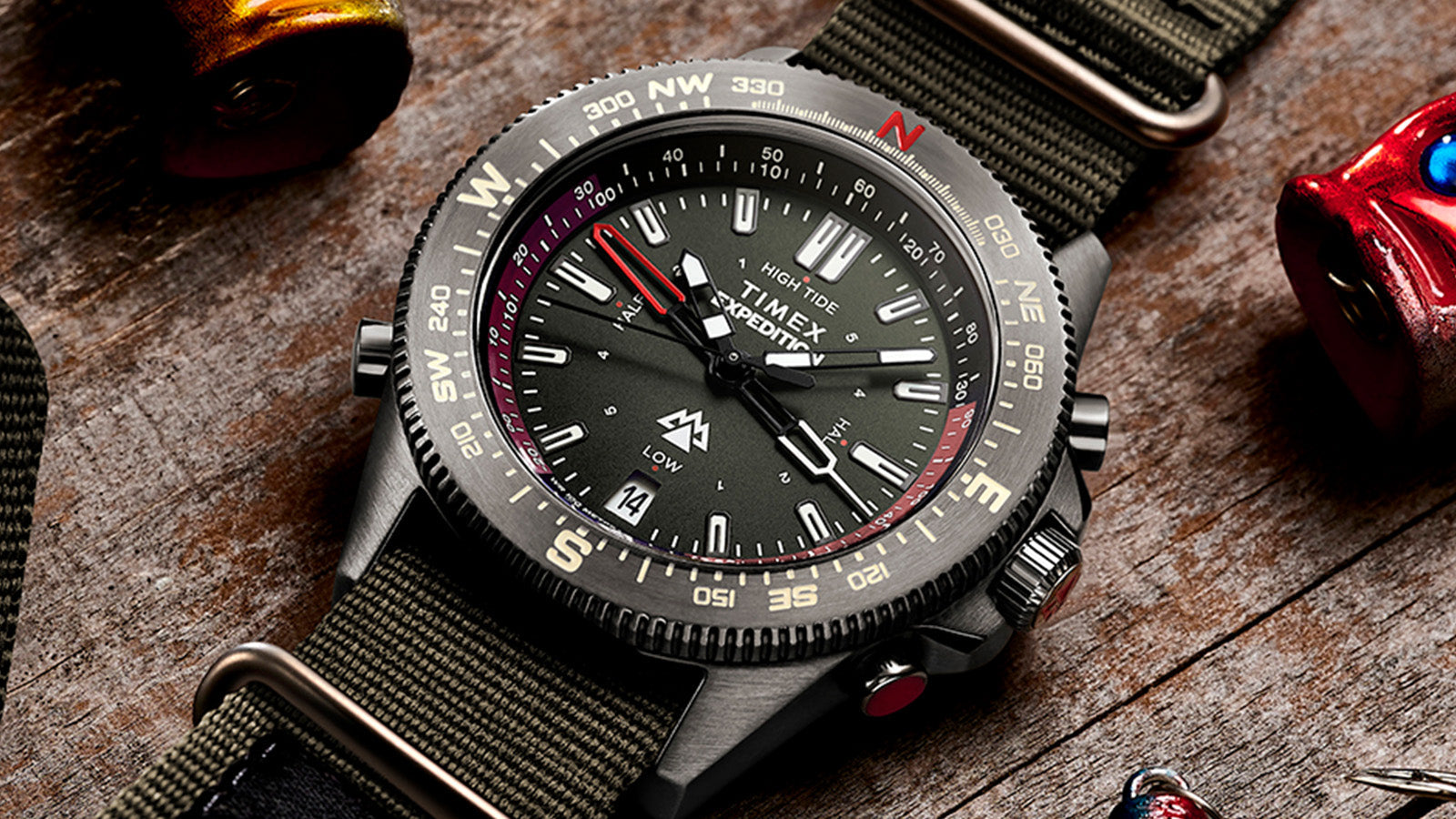 Close-up of the Timex Expedition Tide-Temp-Compass laid on wood with fishing lures, showcasing detailed dial markers, tide indicator, and rugged case design.