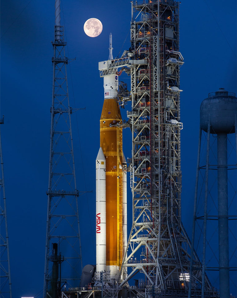 NASA Artemis rocket on a launchpad at night with the full moon in the sky, lit by floodlights and surrounded by launch structures.