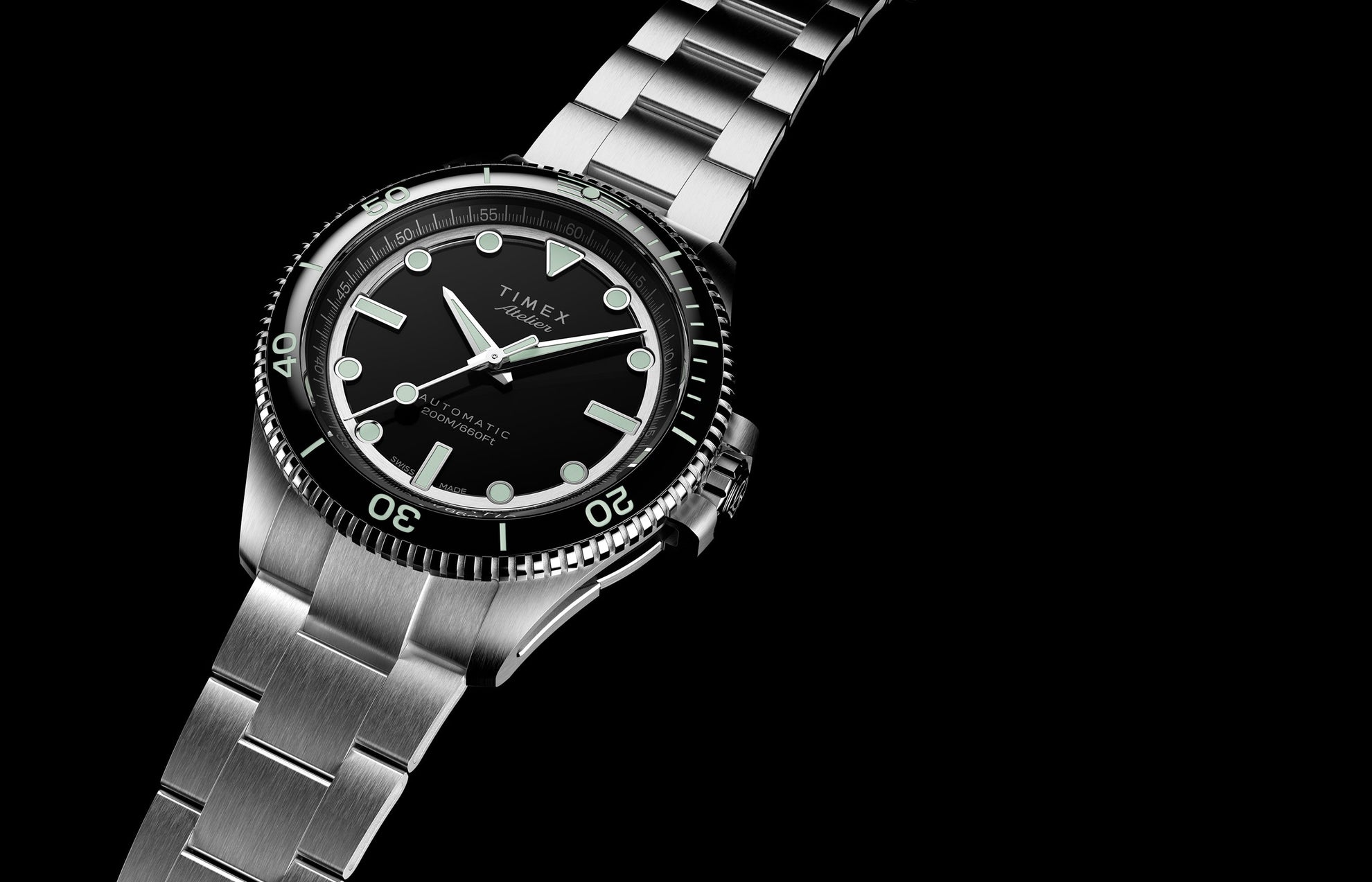 Timex Atelier automatic dive watch in brushed stainless steel with black dial, luminous markers, and unidirectional bezel, angled against a dark background.