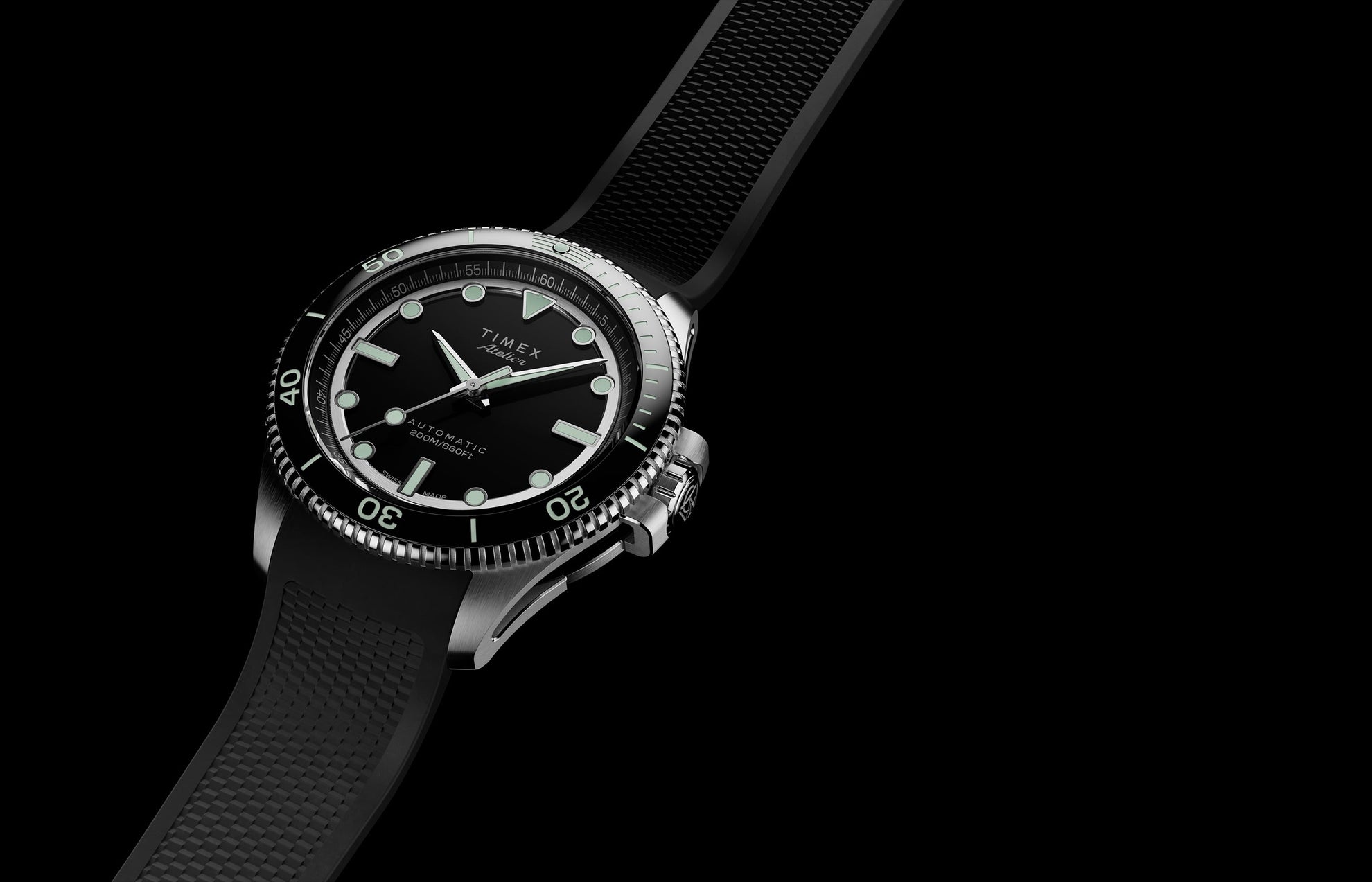 Timex Atelier automatic dive watch with stainless steel case, black dial, luminous markers, and black textured rubber strap on a dark background.