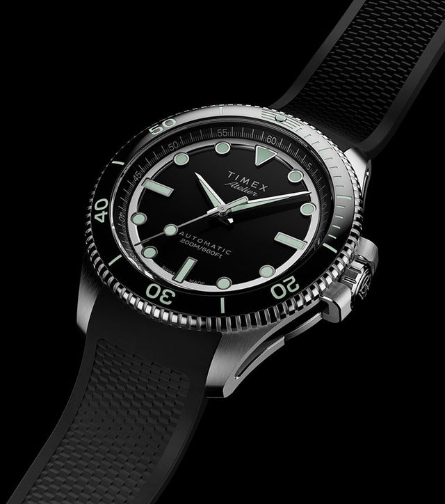 Timex Atelier automatic dive watch with stainless steel case, black dial, luminous markers, and black textured rubber strap on a dark background.