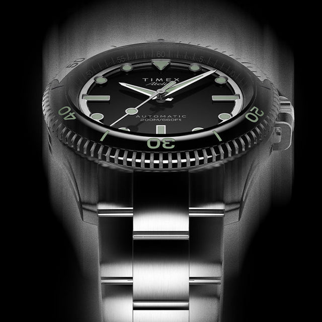 Front view of Timex Atelier automatic dive watch showing black dial, bold luminous indices, stainless steel bracelet, and engraved bezel detail.