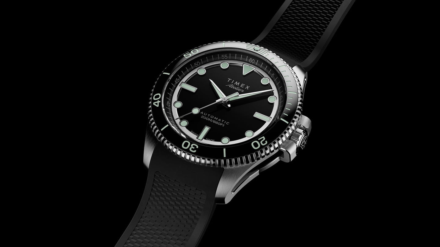 A Timex Atelier automatic dive watch with a black dial, luminous markers, rotating bezel, and a textured black rubber strap, shown in a close-up angled view against a black background.