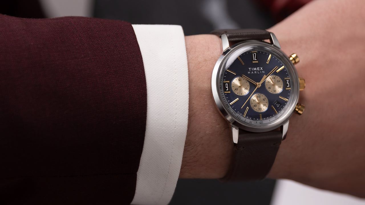 Timex Marlin Chronograph Tachymeter with navy blue dial, gold subdials, and brown leather strap displayed on blue background.
