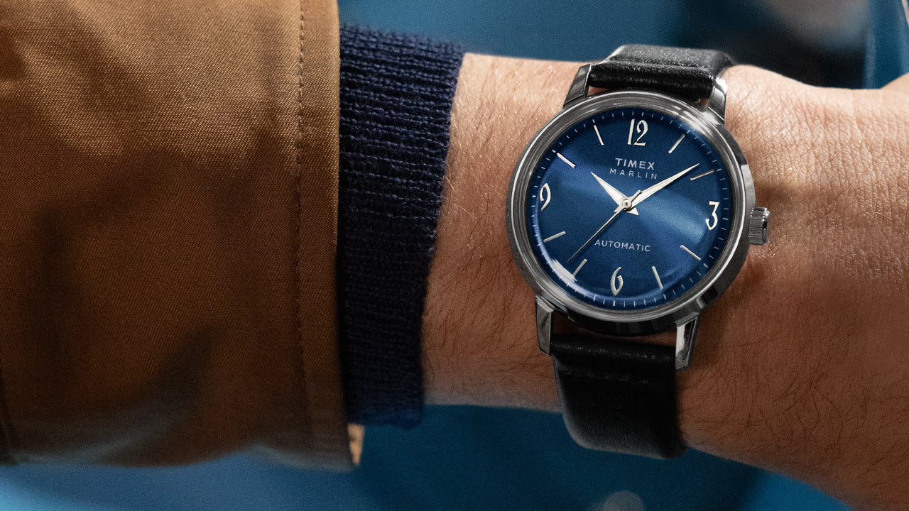 Timex Marlin Automatic watch with blue dial and silver-tone markers worn on wrist, paired with a brown jacket and navy knit cuff.