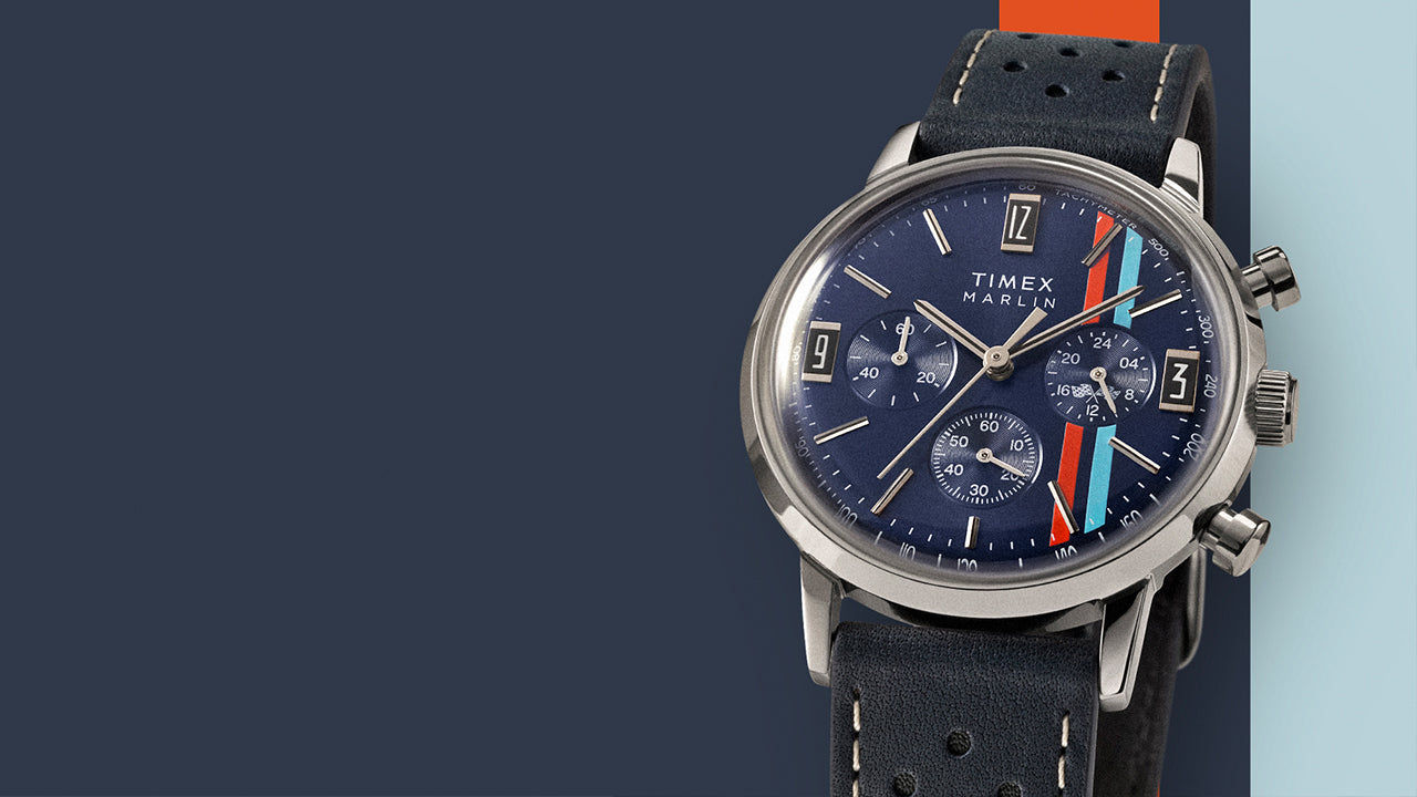 Timex Marlin Chronograph Tachymeter with blue dial, red and light blue racing stripes, and perforated leather strap displayed on a color-blocked blue and orange background.