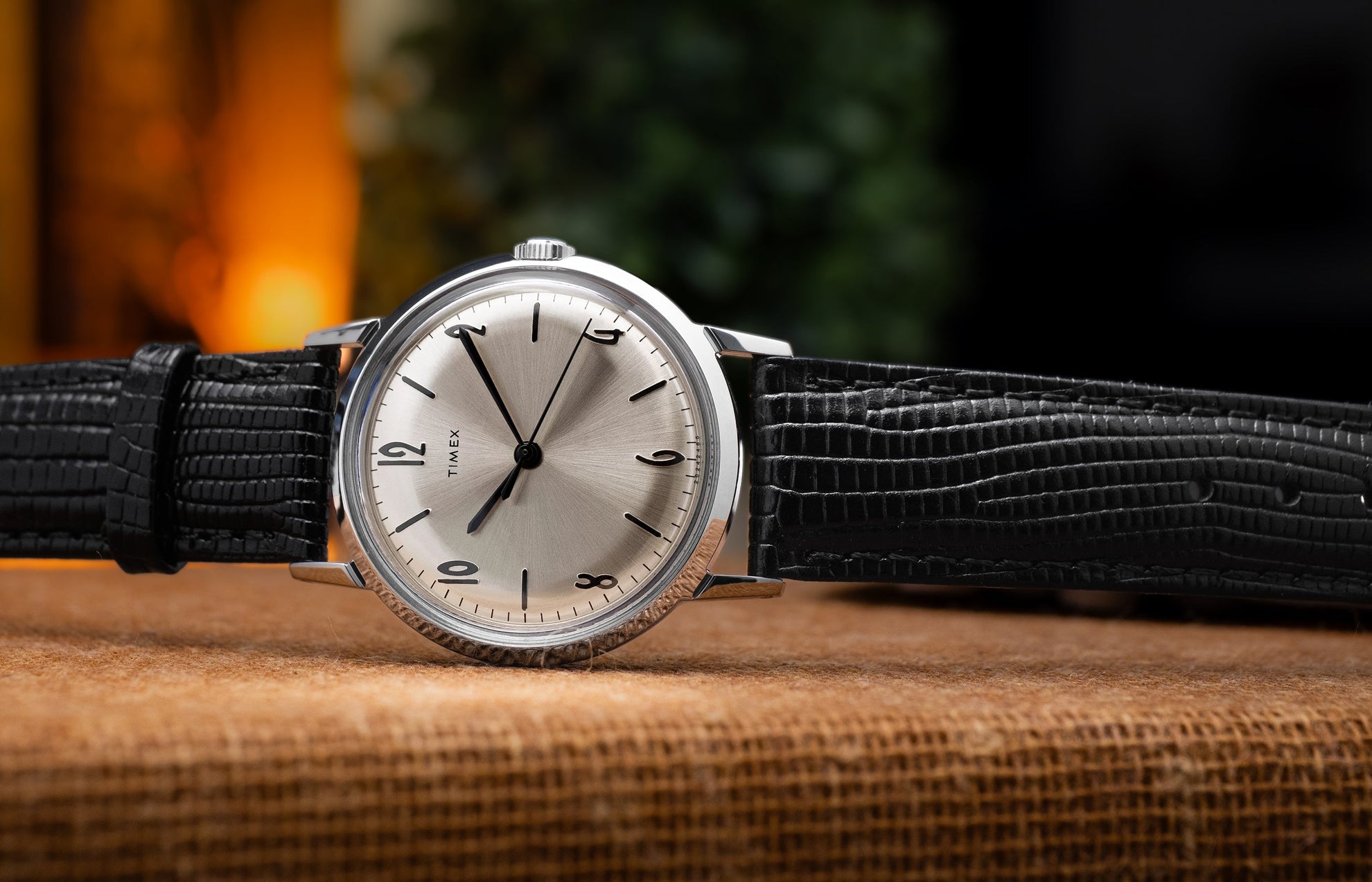 Timex Marlin Hand-Wound watch with a silver sunburst dial, black hour markers, and black leather strap resting on a brown fabric surface, warm background lighting.