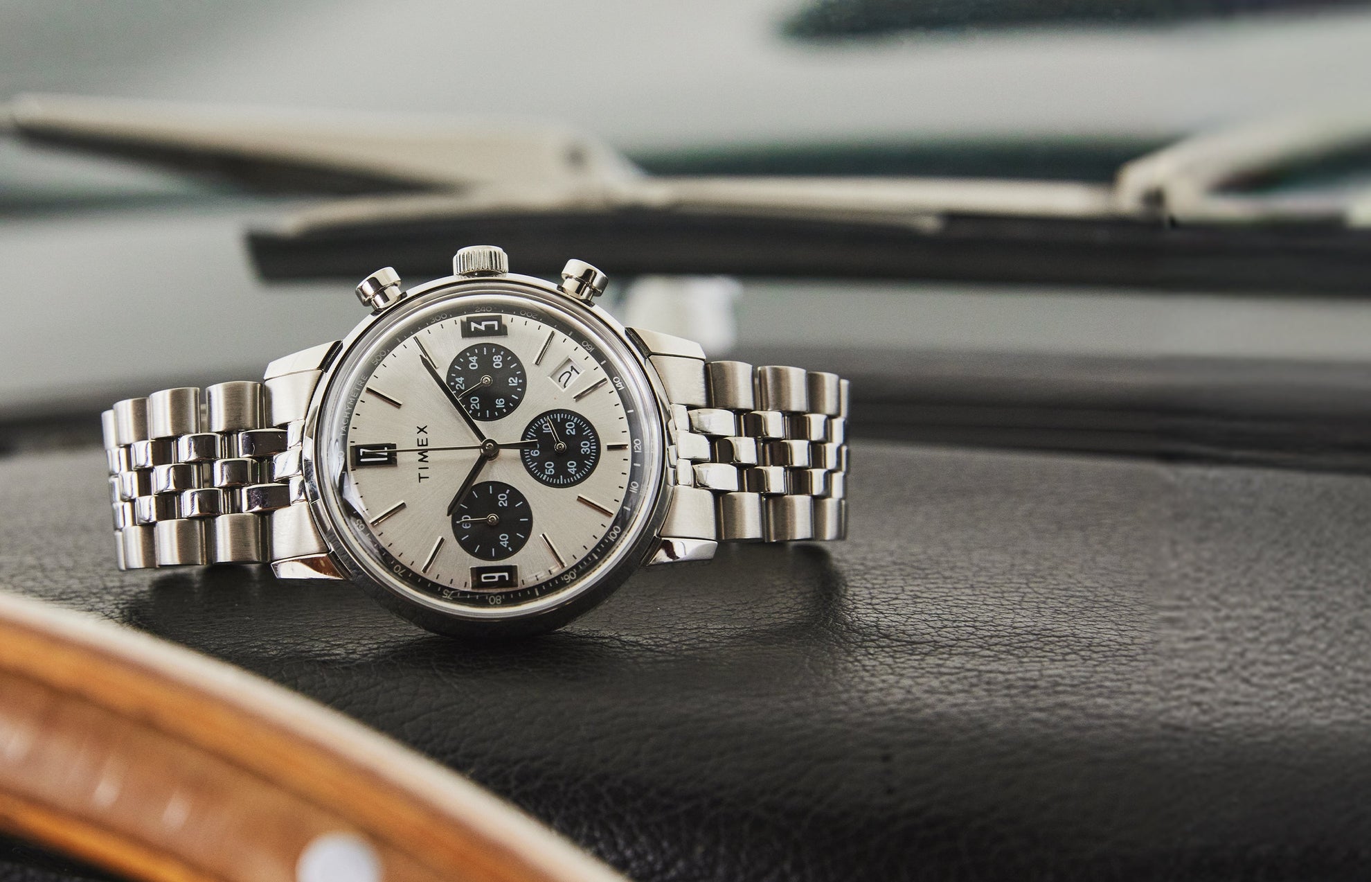 Timex Marlin Chronograph Tachymeter with silver dial, black subdials, and stainless-steel bracelet resting on black leather car dashboard.