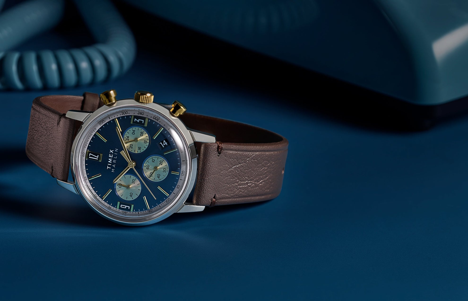 Timex Marlin Chronograph Tachymeter with blue dial and gold accents worn on wrist, styled with burgundy suit jacket and white cuff.