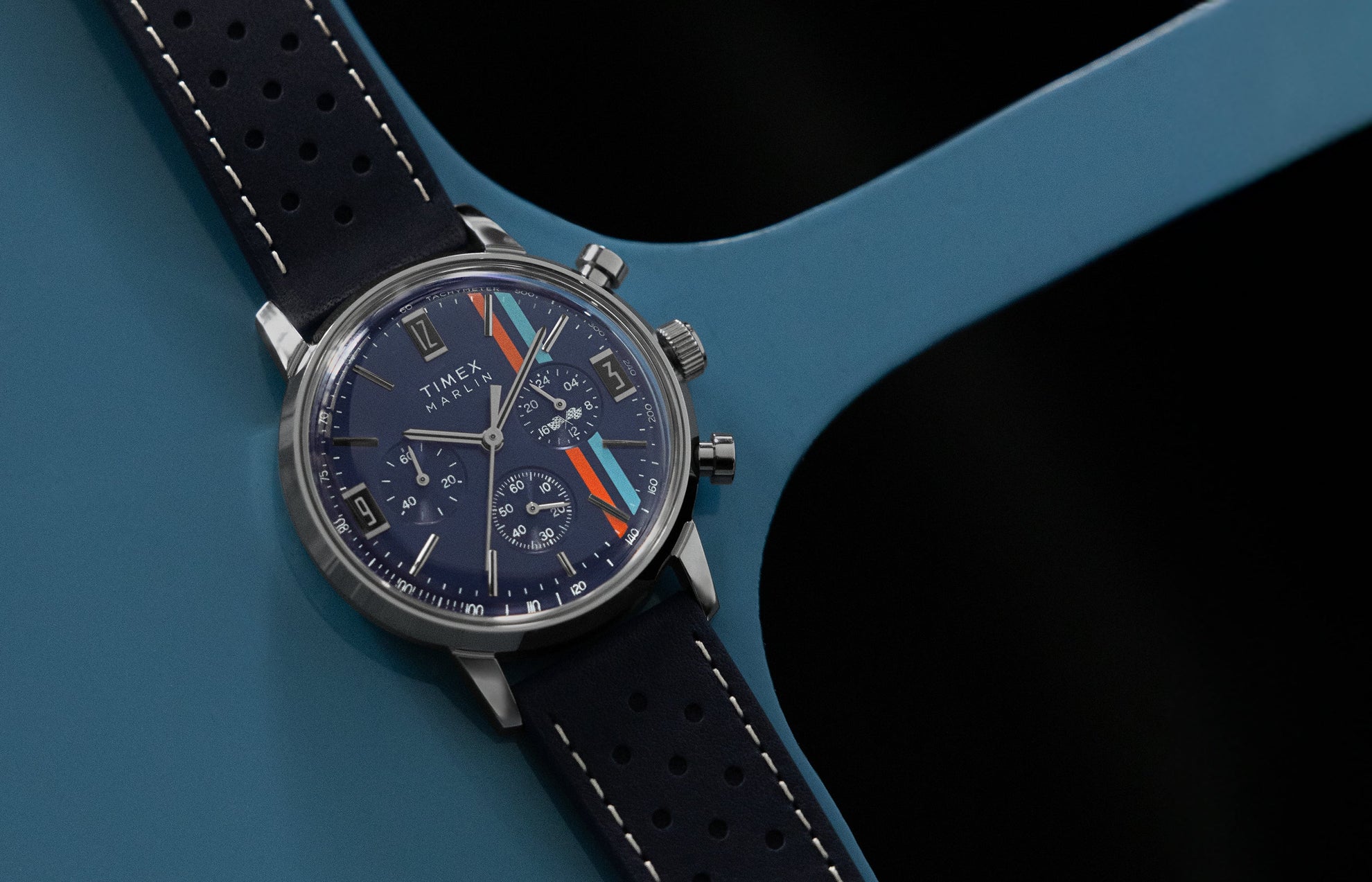 Timex Marlin Chronograph Tachymeter with navy dial, multi-color racing stripes, and perforated blue leather strap resting on a blue metal surface.