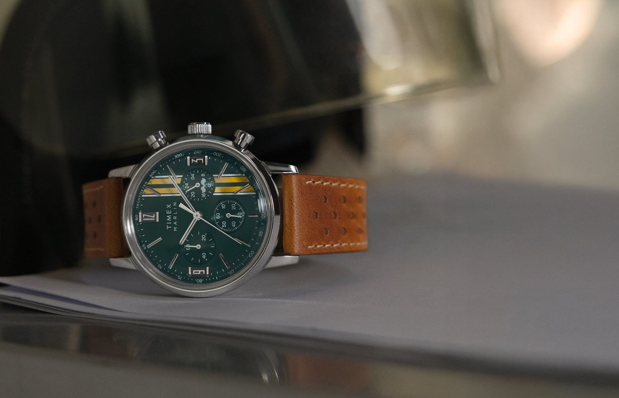 Timex Marlin Chronograph Tachymeter with green dial, yellow racing stripes, and brown perforated leather strap resting on a metallic surface.