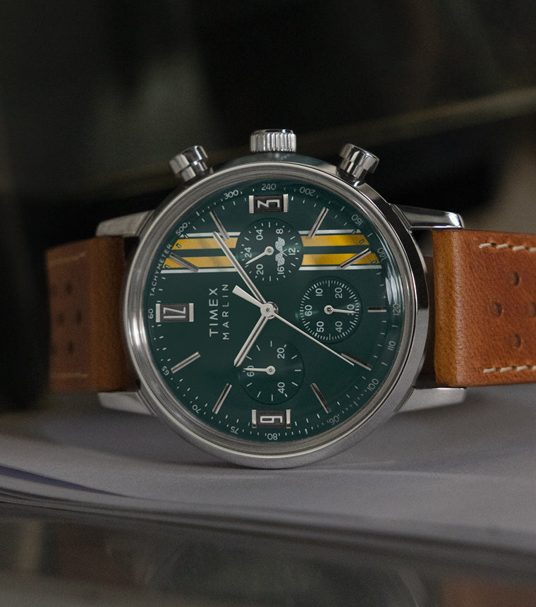 Timex Marlin Chronograph Tachymeter with green dial, yellow racing stripes, and brown perforated leather strap resting on a metallic surface.