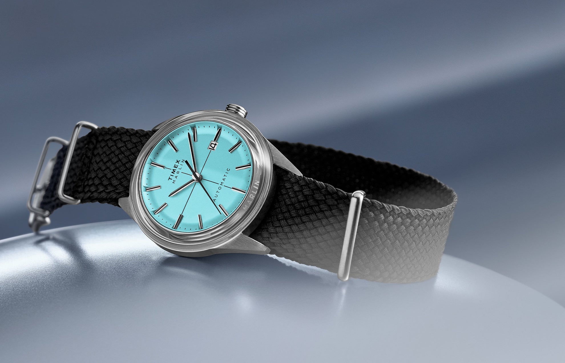 Timex Marlin Jet Automatic with aqua blue dial, stainless steel case, and black woven strap displayed on a metallic blue surface.