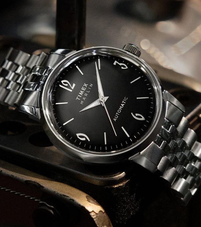 Timex Marlin Automatic with black dial and silver stainless-steel bracelet resting on a vintage metal surface.