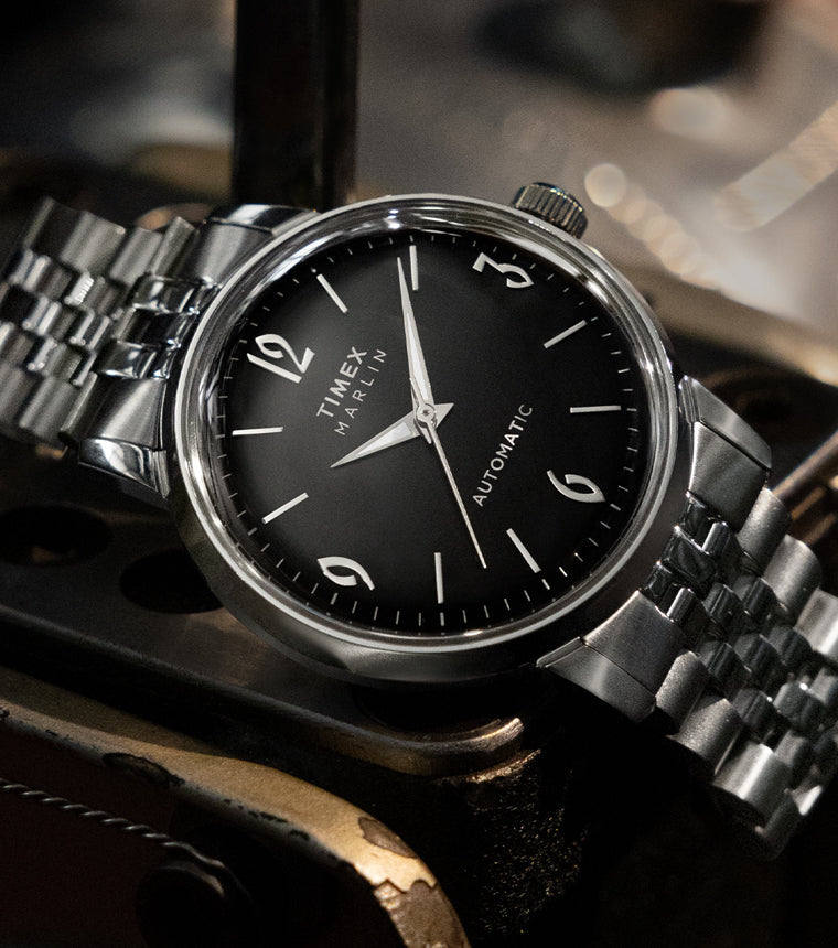 Timex Marlin Automatic with black dial and silver stainless-steel bracelet resting on a vintage metal surface.