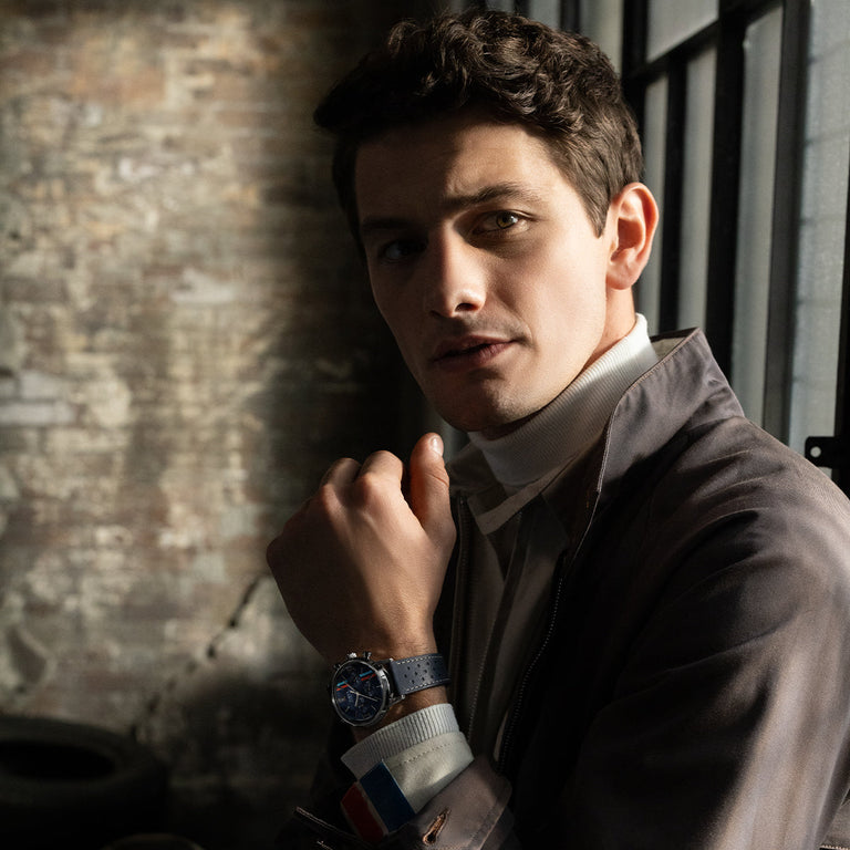 Model wearing Timex Marlin Chronograph Tachymeter with blue dial and racing stripes, in soft window light with exposed brick background.