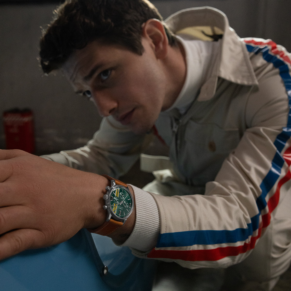 Model in a cream racing jacket with red and blue stripes, wearing the Timex Marlin Chronograph Tachymeter with green dial and brown leather strap.