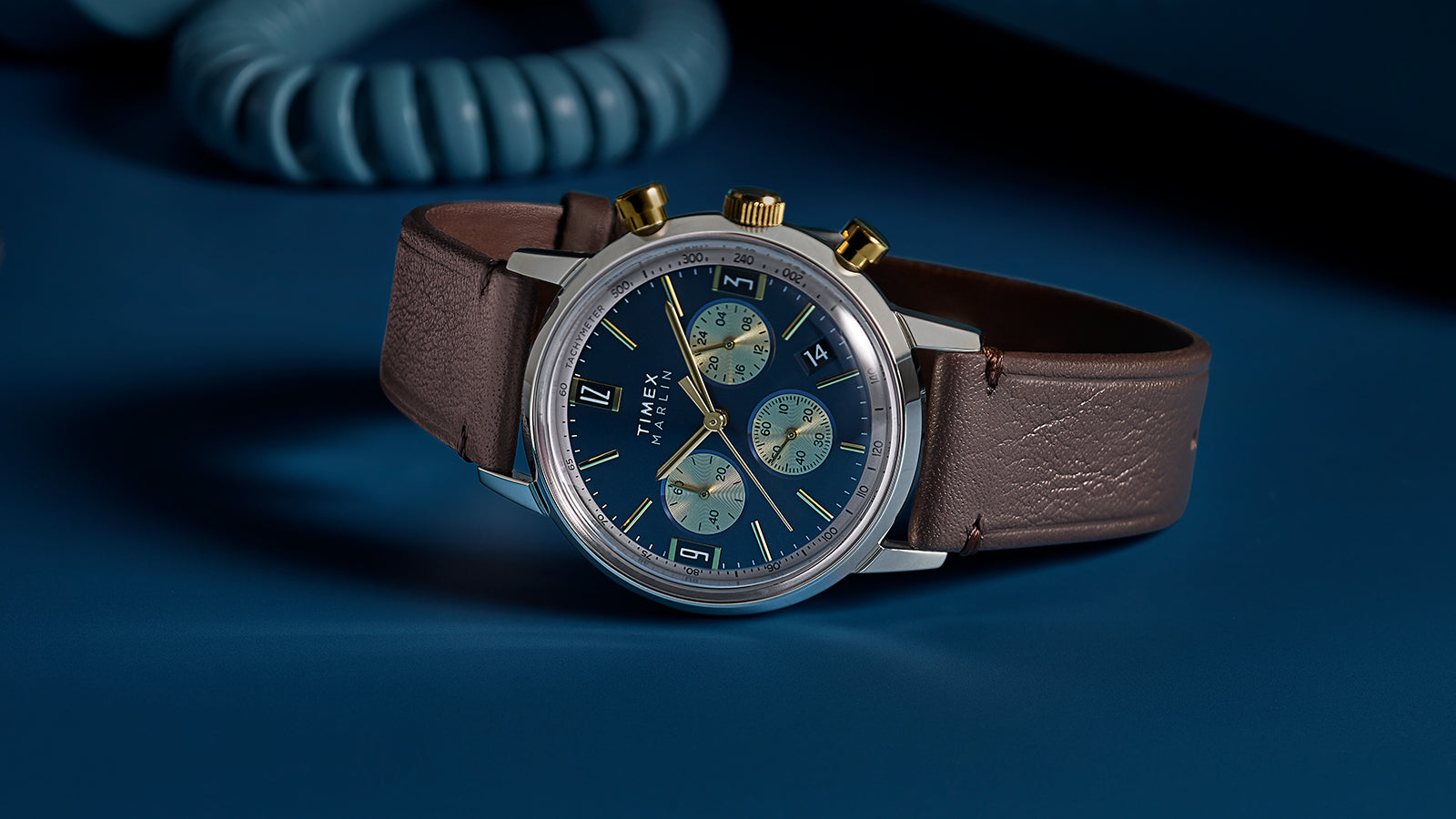 Timex Marlin Chronograph Tachymeter with blue dial and gold accents worn on wrist, styled with burgundy suit jacket and white cuff.