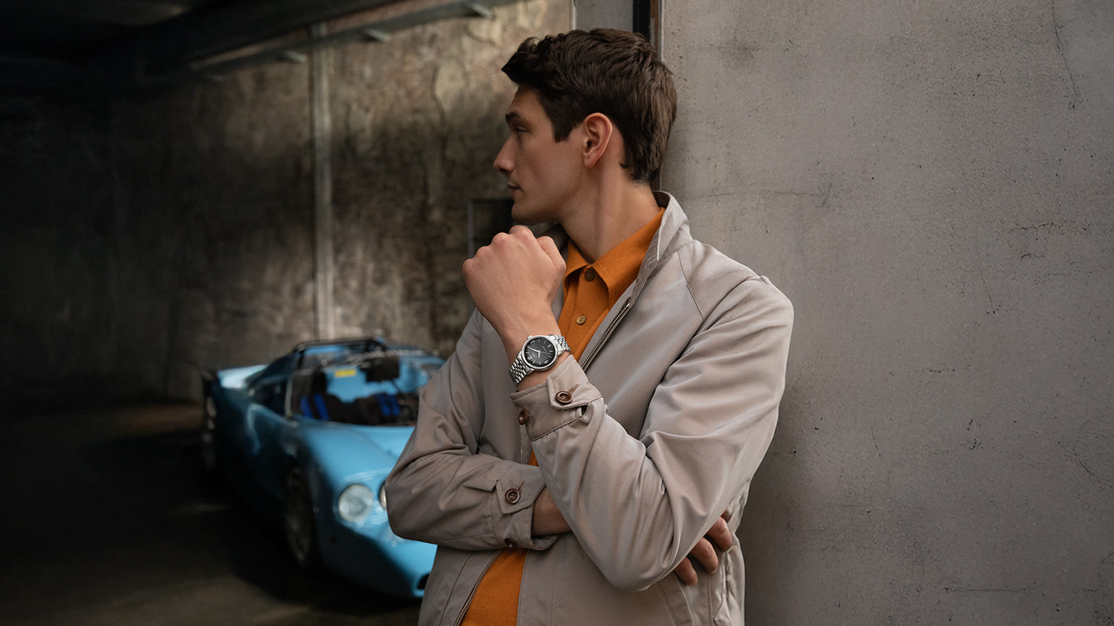 Model wearing Timex Marlin Automatic with black dial and stainless-steel bracelet, styled in beige jacket and orange shirt, standing beside a blue vintage car.