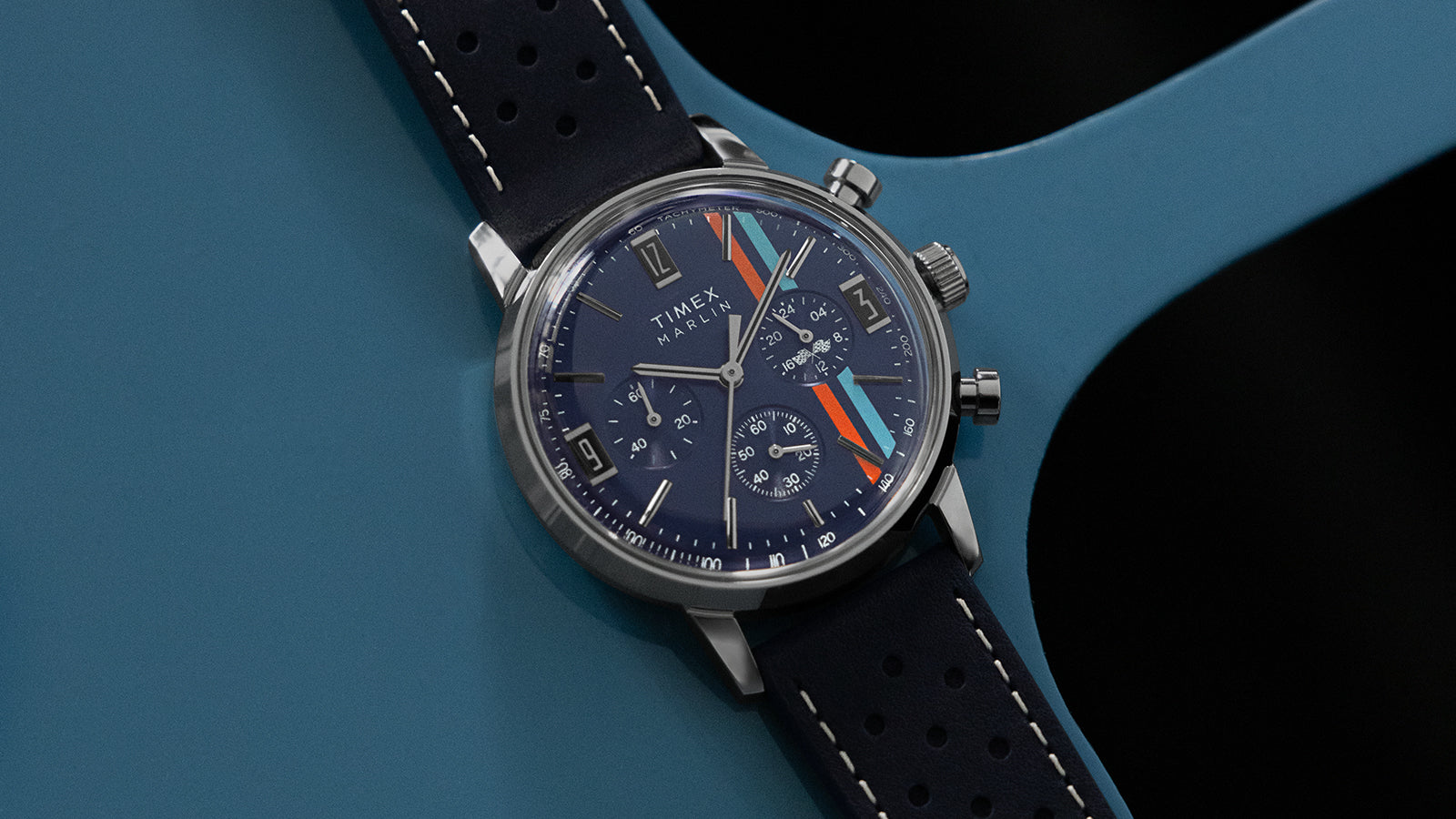 Timex Marlin Chronograph Tachymeter with navy dial, multi-color racing stripes, and perforated blue leather strap resting on a blue metal surface.