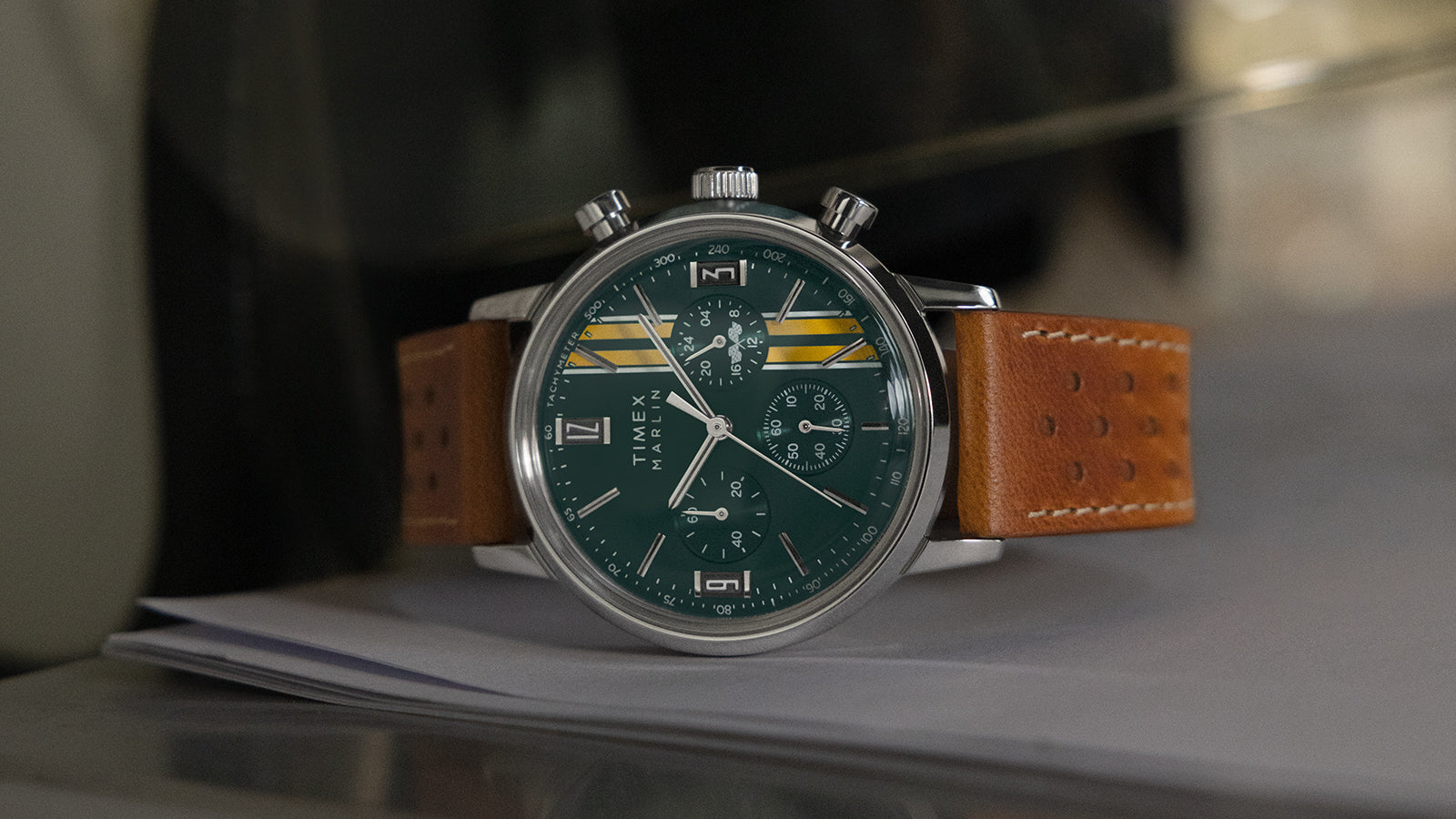 Timex Marlin Chronograph Tachymeter with green dial, yellow racing stripes, and brown perforated leather strap resting on a metallic surface.