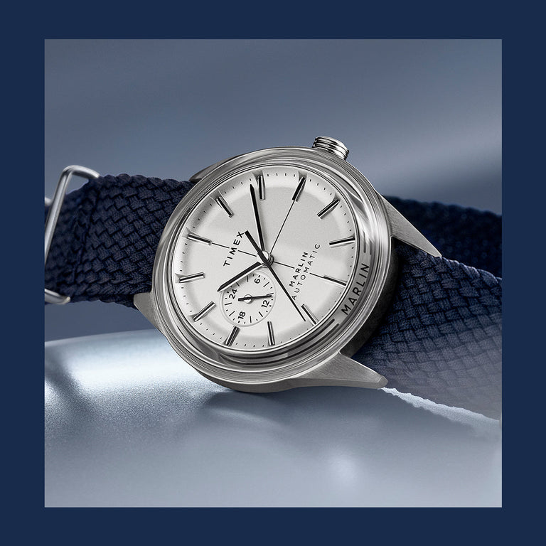 Timex Marlin Automatic watch featuring a silver dial, subdial at 9 o’clock, and navy woven strap, shown against a cool metallic background.