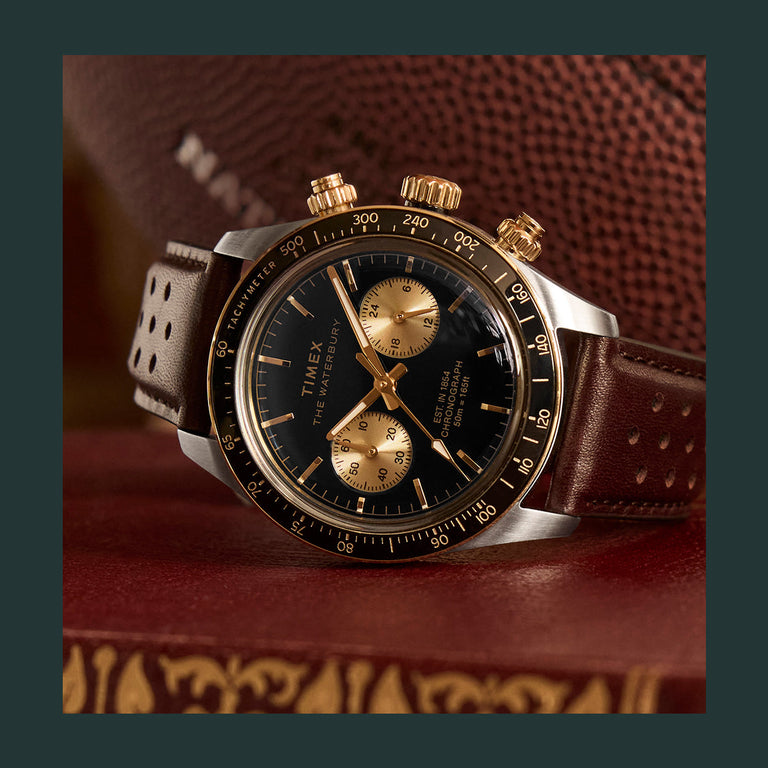 Timex Waterbury Chronograph with a black dial, gold subdials, and brown perforated leather strap, photographed on a vintage football background.