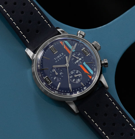 Timex Marlin chronograph watch with a navy blue dial, silver case, and perforated blue leather strap featuring orange racing stripes, photographed on a blue surface.