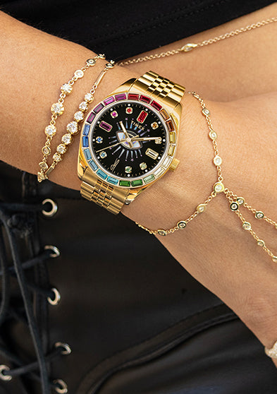 Timex x Jacquie Aiche “Cosmic Nights” watch with a gold-tone case and bracelet featuring multicolored gemstone accents. The black dial displays crystal hour markers and a radiant “all-seeing eye” motif at the center, blending celestial and luxury design elements.