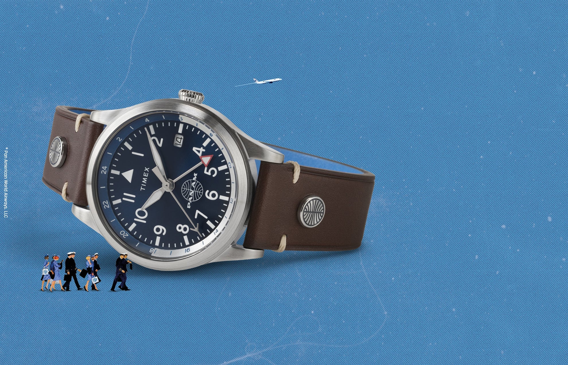 Timex x Pan Am® Waterbury GMT watch with a deep blue dial, silver stainless-steel case, and brown leather strap featuring Pan Am-branded rivets. Displayed on a sky-blue background with miniature Pan Am flight crew illustrations and an airplane in the distance, evoking classic aviation style.