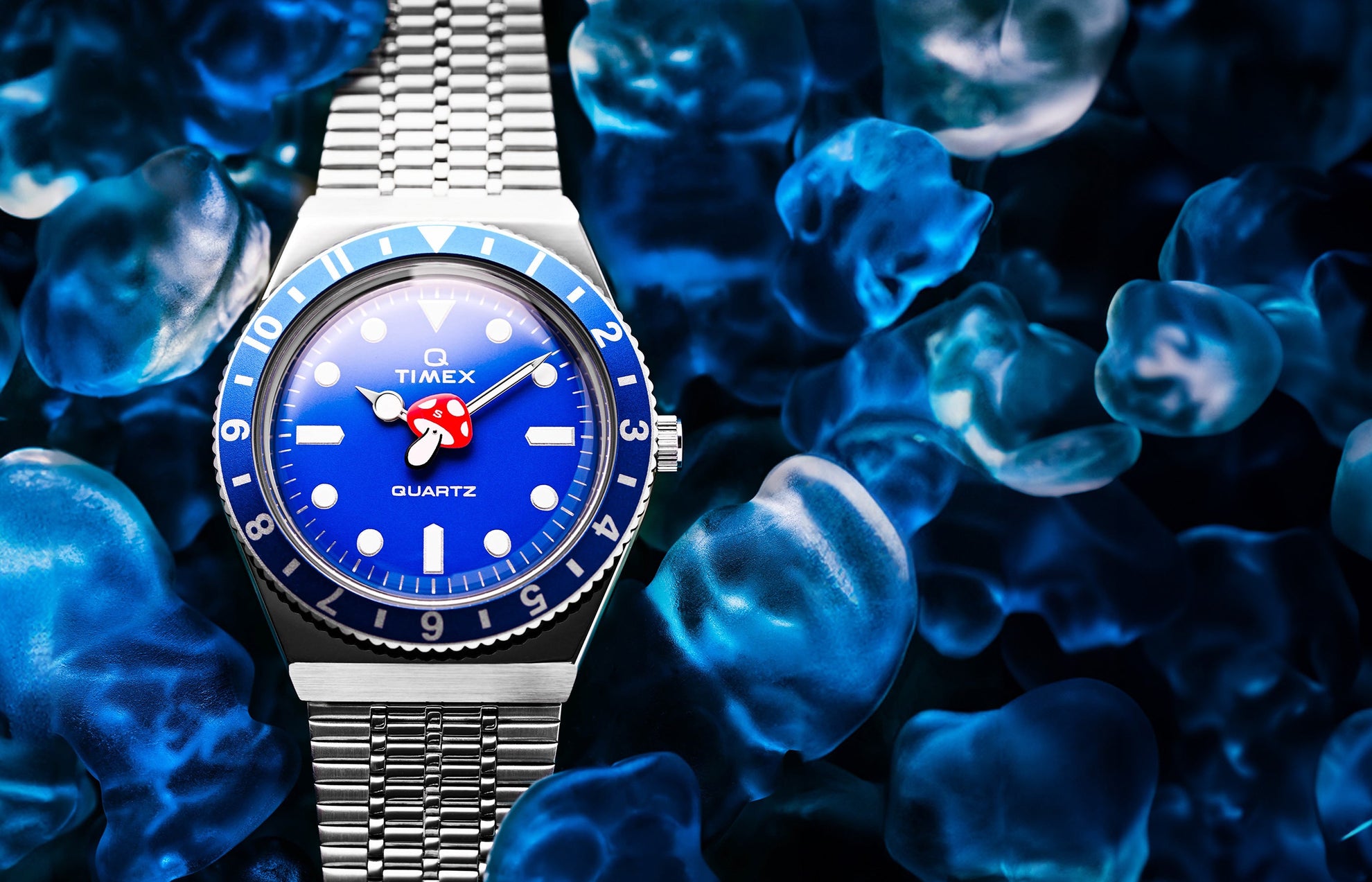 A Q Timex watch with a bright blue dial and red mushroom-shaped seconds hand, set against a background of translucent blue gummy candies. The watch has a stainless-steel bracelet and a blue bezel.