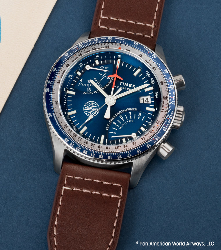 Close-up of the Timex x Pan Am® Fly-Back Chronograph watch with a blue dial, silver-tone case, and brown leather strap. The watch face features Pan Am’s globe logo, multiple sub-dials, and a rotating bezel with white and blue detailing, placed on a blue background beside vintage travel documents.