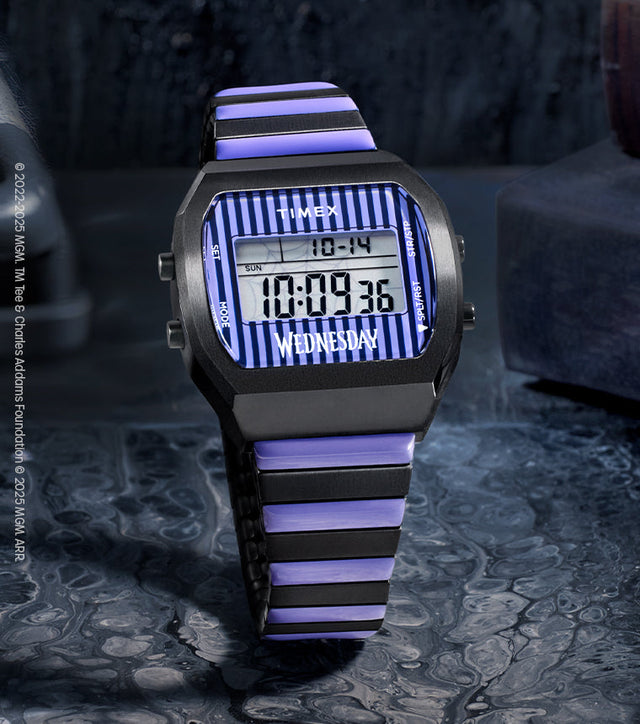 Timex x Wednesday T80 digital watch featuring a black and purple striped design on the face and bracelet. The watch is displayed on a dark, moody surface beside vintage books, evoking a gothic aesthetic inspired by Wednesday Addams.