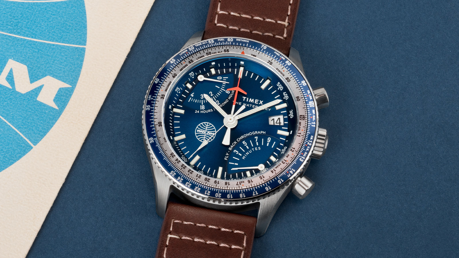 Detailed top view of the Timex x Pan Am® Fly-Back Chronograph showing its blue sunray dial, chronograph sub-dials, and Pan Am globe emblem. The silver bezel features aviation-style markings, and the brown stitched leather strap adds a vintage pilot aesthetic.