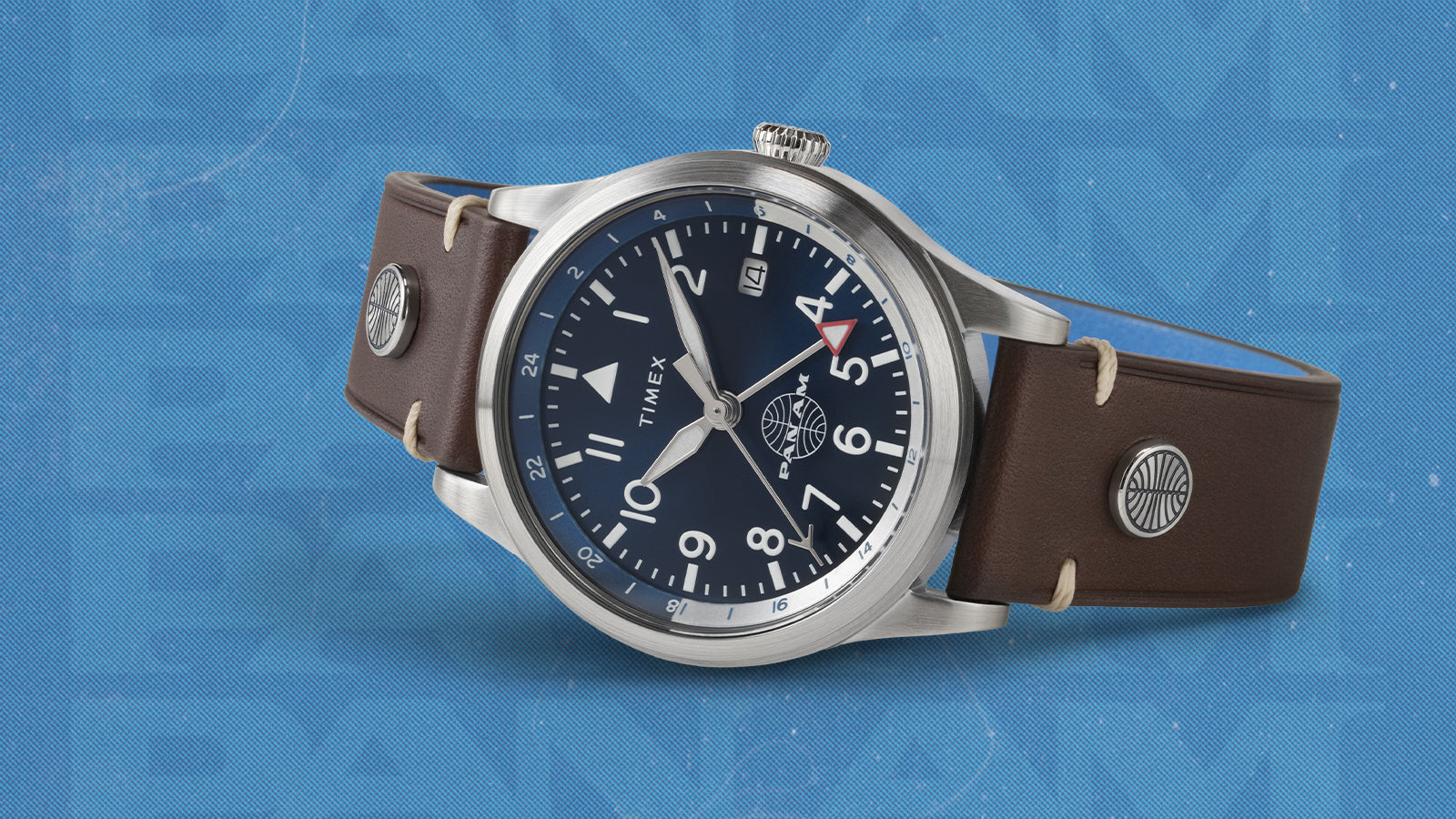 Close-up angled view of the Timex x Pan Am® Waterbury GMT showing its navy dial, silver markers, and red-tipped GMT hand. The Pan Am globe logo sits above 6 o’clock, and the brown stitched leather strap adds a refined travel aesthetic against a blue textured background.