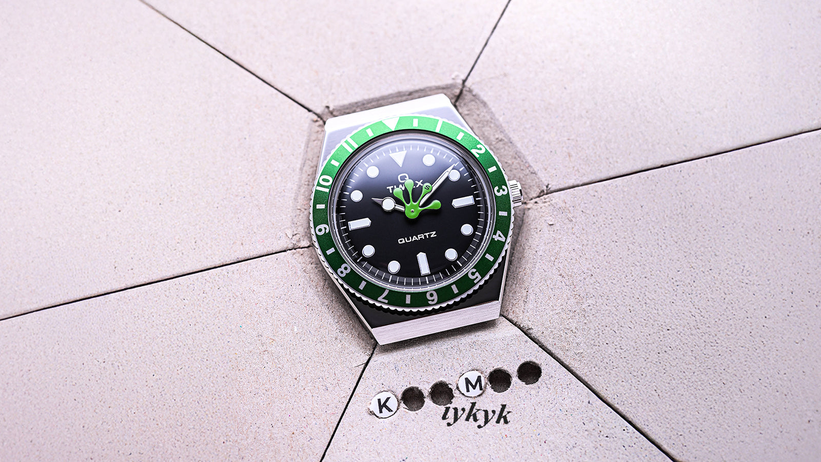 Overhead view of the Timex x seconde/seconde/ Q Timex watch with a green bezel and playful frog foot hand placed on a gray geometric surface etched with “KM tykyk” details, blending humor with horology.